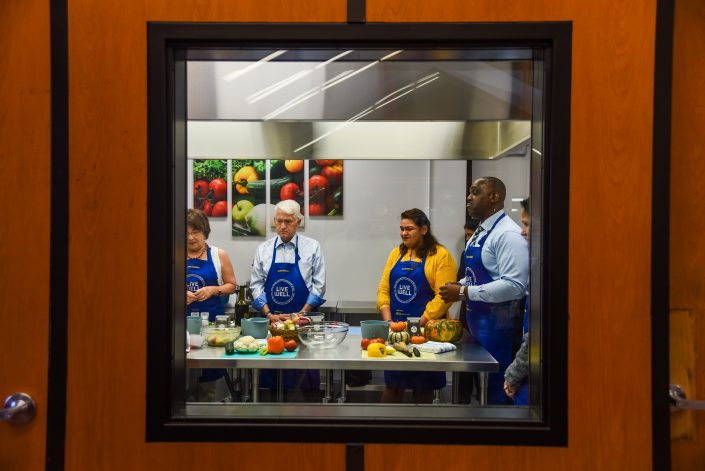 Photographed through a window, two women and two men stand around a kitchen table with ingredients.