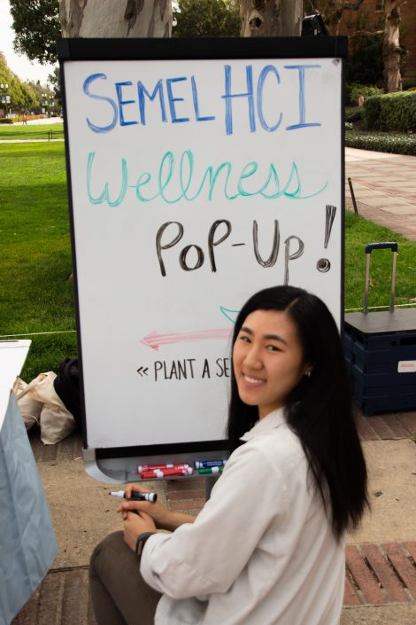 Woman in white smiles in front of "Wellness Pop-Up" poster.